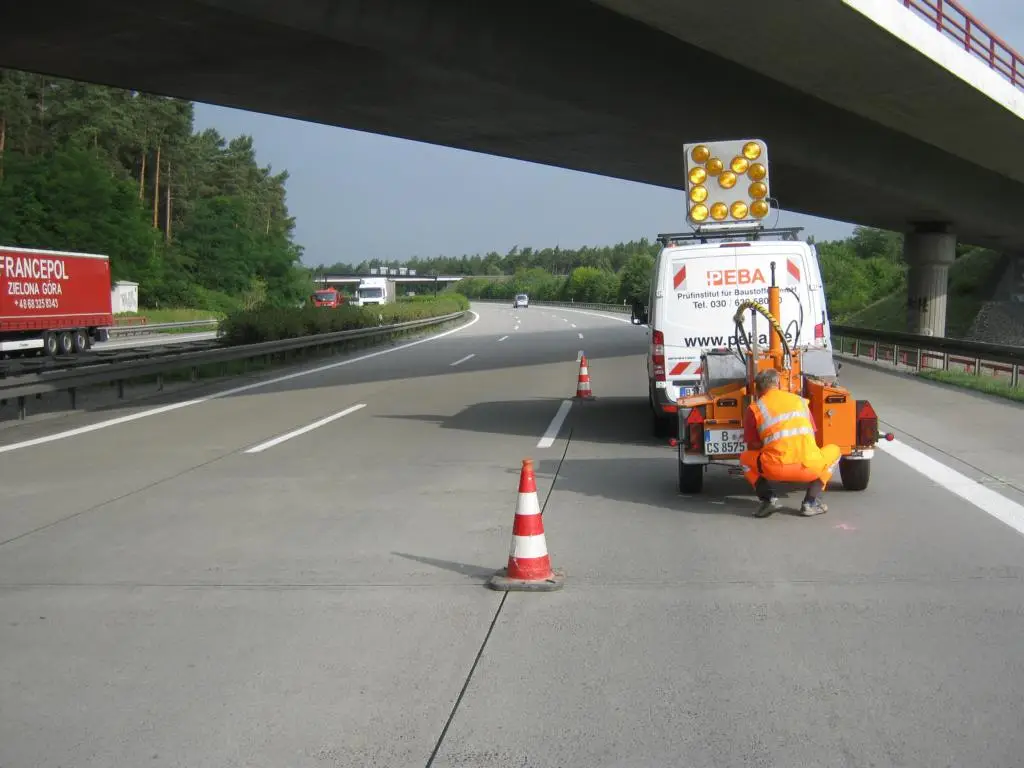 Bauarbeiter in orangefarbener Sicherheitskleidung bei Prüfungen auf der Bundesautobahn A2, PEBA Fahrzeug mit Warnleuchten und Verkehrsabsperrungen im Hintergrund.