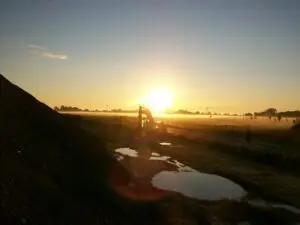 Bauarbeiten am Militärflugplatz Holzdorf bei Sonnenaufgang, Baustellenfahrzeug im Vordergrund, nasse Bodenflächen, umgebende Nebel- und Landschaftselemente.